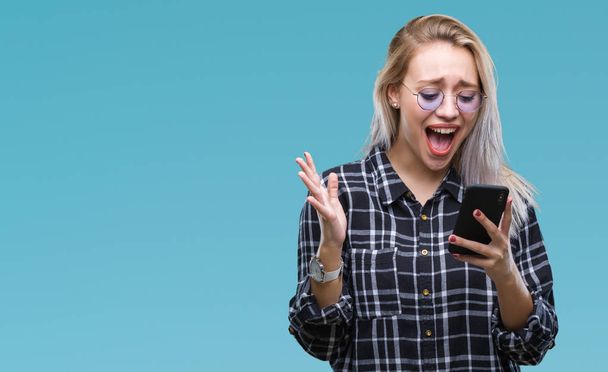 Fiatal szőke nő SMS üzenetet használ smartphone felső elszigetelt háttér nagyon boldog és izgatott, győztes kifejezés ünnepli győzelem nagy mosollyal sikoltozik, és kezet emelt - Fotó, kép
