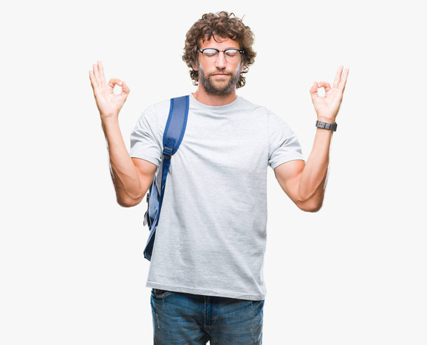 Hombre estudiante hispano guapo con mochila y gafas sobre fondo aislado relajarse y sonreír con los ojos cerrados haciendo gesto de meditación con los dedos. Concepto de yoga
. - Foto, Imagen