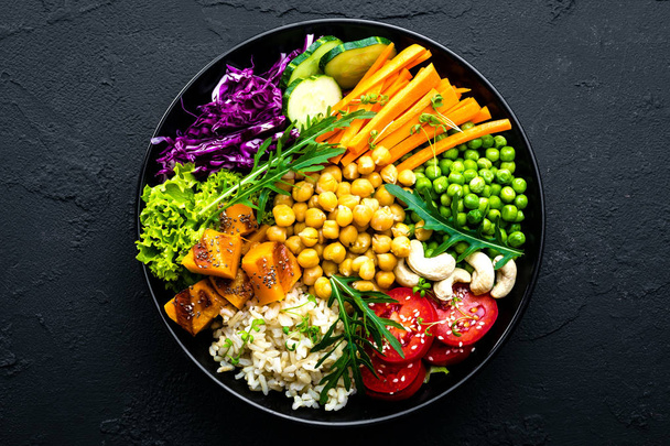 Bowl dish with brown rice, cucumber, tomato, green peas, red cabbage, chickpea, fresh lettuce salad and cashew nuts. Healthy balanced eating - Photo, Image