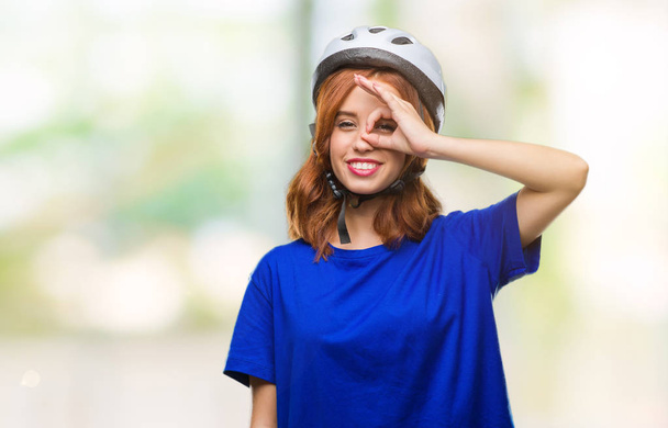 Jovem mulher bonita vestindo capacete ciclista sobre fundo isolado fazendo gesto ok com a mão sorrindo, olho olhando através dos dedos com rosto feliz
. - Foto, Imagem
