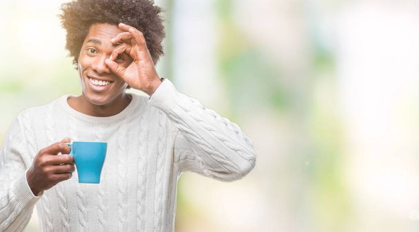 Afro-amerikai ember iszik kávét elszigetelt háttérhez képest boldog arc mosolyog, csinál a kéz-szem nézegette ujjak ok jele - Fotó, kép