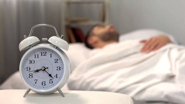 Male sleeping in bed, white alarm clock standing on table morning, biorhythm - Photo, Image