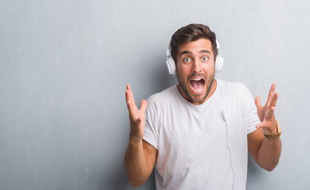 Knappe jongeman over grijs grunge muur dragen koptelefoon luisteren naar muziek erg blij en opgewonden, winnaar expressie vieren overwinning schreeuwen met grote glimlach en getogen handen - Foto, afbeelding