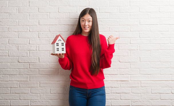 Young Chinese real state agent woman over brick wall holding house pointing and showing with thumb up to the side with happy face smiling - Foto, immagini