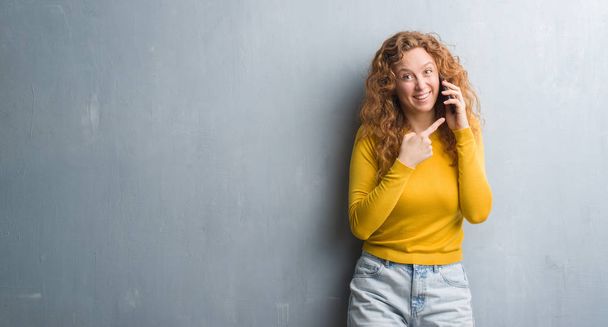 Fiatal vöröshajú nő beszél a telefonon nagyon boldog, rámutatva a kéz és az ujjak szürke grunge falon - Fotó, kép