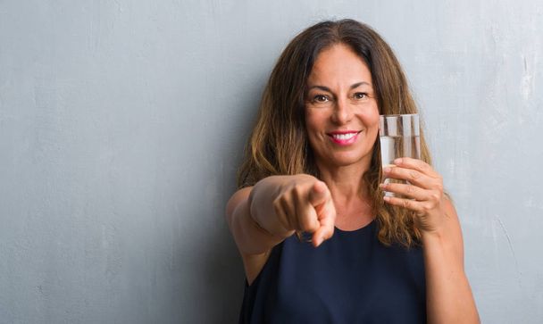 Middelbare leeftijd Spaanse vrouw drinken glas water met de vinger te wijzen naar de camera en u, hand ondertekenen, positieve en zelfverzekerd gebaar van de voorkant - Foto, afbeelding