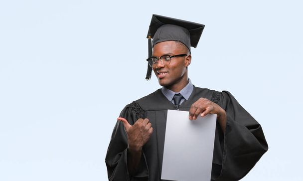 Young graduated african american man holding blank paper degree over isolated background pointing and showing with thumb up to the side with happy face smiling - Фото, изображение
