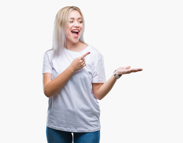 Jonge blonde vrouw op geïsoleerde achtergrond verbaasd en lachend naar de camera tijdens het presenteren met hand en met de vinger te wijzen. - Foto, afbeelding