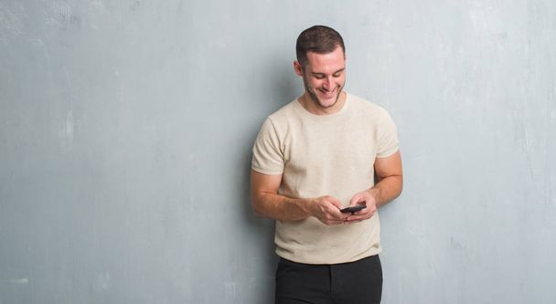 Jeune homme caucasien sur un mur gris grunge texter un message à l'aide d'un smartphone avec un visage heureux debout et souriant avec un sourire confiant montrant les dents
 - Photo, image