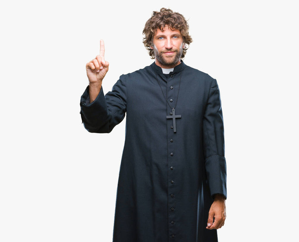 Hezký hispánský katolický kněz muž nad izolované pozadí zobrazení a směřující nahoru s prstem číslo jedna při úsměvu a spokojené. - Fotografie, Obrázek
