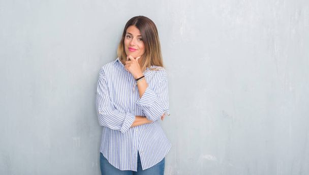 Jonge volwassen vrouw over grijs grunge muur dragen mode zakelijke outfit op zoek vertrouwen op de camera met glimlach met gekruiste armen en hand op kin aan de orde gesteld. Positief denken. - Foto, afbeelding