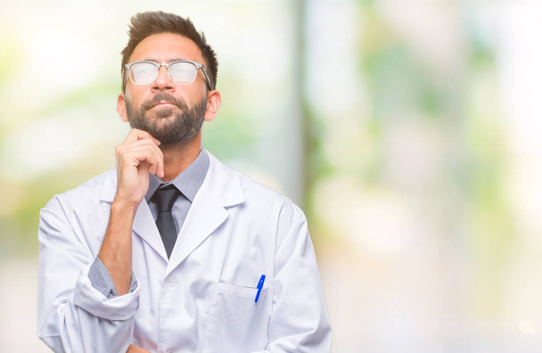 Adult hispanic scientist or doctor man wearing white coat over isolated background with hand on chin thinking about question, pensive expression. Smiling with thoughtful face. Doubt concept. - Photo, Image