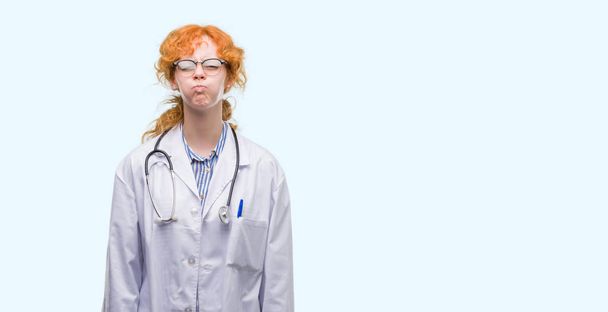 Mladá zrzka žena nosí uniformu doktor nafoukla tváře s legrační obličej. Ústa naplněny vzduchem, šílený výraz. - Fotografie, Obrázek