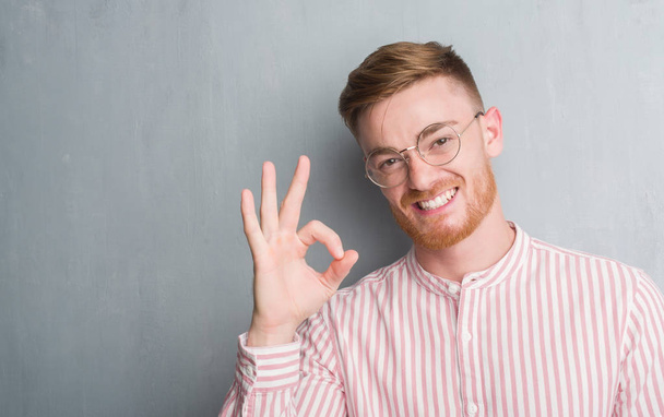 Jonge roodharige man over grijs grunge muur doen ok teken met vingers, uitstekende symbool - Foto, afbeelding