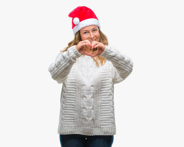 Mujer hispana mayor de mediana edad con sombrero de Navidad sobre fondo aislado sonriendo en el amor mostrando el símbolo del corazón y la forma con las manos. Concepto romántico
. - Foto, Imagen