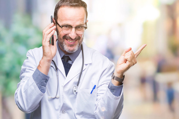 Старший седовласый врач средних лет, разговаривающий на смартфоне на изолированном фоне, очень доволен, указывая рукой и пальцем в сторону.
 - Фото, изображение