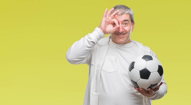 Bello uomo anziano che tiene palla da calcio su sfondo isolato con volto felice sorridente facendo ok segno con mano sugli occhi guardando attraverso le dita
 - Foto, immagini