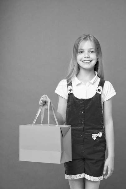 Little child with shopping bag on violet background. Happy girl smile with paper bag. Kid shopper in fashion jumpsuit. Holidays and celebration. Satisfied with her shopping. sale and black friday - Foto, Imagen