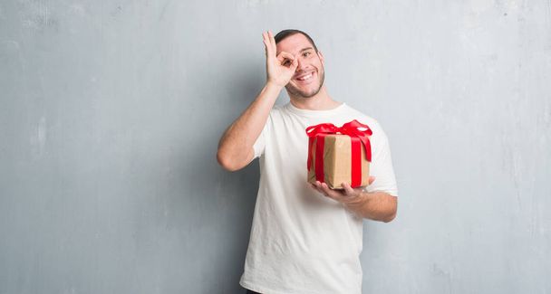 Homem caucasiano jovem sobre parede grunge cinza segurando um presente com o rosto feliz sorrindo fazendo ok sinal com a mão no olho olhando através dos dedos
 - Foto, Imagem