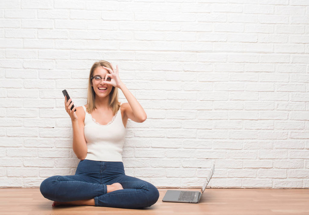 Красивая молодая женщина сидит на полу смс с помощью смартфона и ноутбука с счастливым лицом улыбается делает хорошо знак с рукой на глазу, глядя сквозь пальцы
 - Фото, изображение