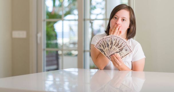 Down syndrome woman at home holding dollars cover mouth with hand shocked with shame for mistake, expression of fear, scared in silence, secret concept - Valokuva, kuva