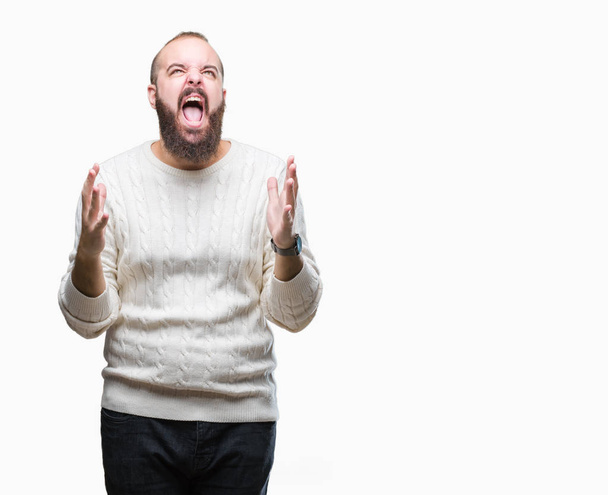 Молодой кавказский хипстер в зимнем свитере на изолированном фоне, сумасшедший, кричащий и кричащий с агрессивным выражением лица и поднятыми руками. Концепция разочарования
. - Фото, изображение