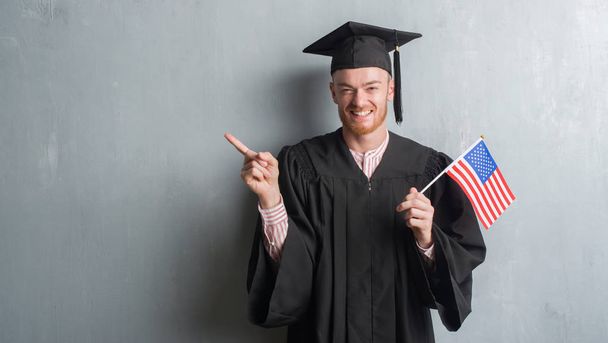 Jovem ruiva homem sobre parede grunge cinza vestindo uniforme graduado segurando bandeira americana muito feliz apontando com a mão e o dedo para o lado
 - Foto, Imagem
