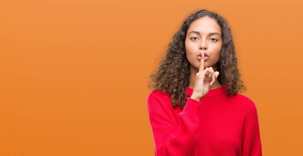 junge hispanische Frau in rotem Pullover, die mit dem Finger auf den Lippen um Ruhe bittet. Schweigen und geheimes Konzept. - Foto, Bild