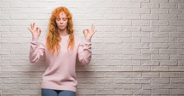 Jonge roodharige vrouw stond over bakstenen muur ontspannen en lachend met ogen gesloten doen meditatie gebaar met vingers. Yoga concept. - Foto, afbeelding