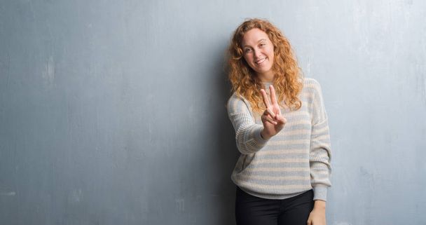 Young redhead woman over grey grunge wall smiling looking to the camera showing fingers doing victory sign. Number two. - Foto, Imagen
