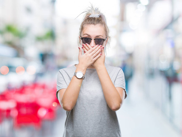 Young blonde woman wearing sunglasses over isolated background shocked covering mouth with hands for mistake. Secret concept. - 写真・画像