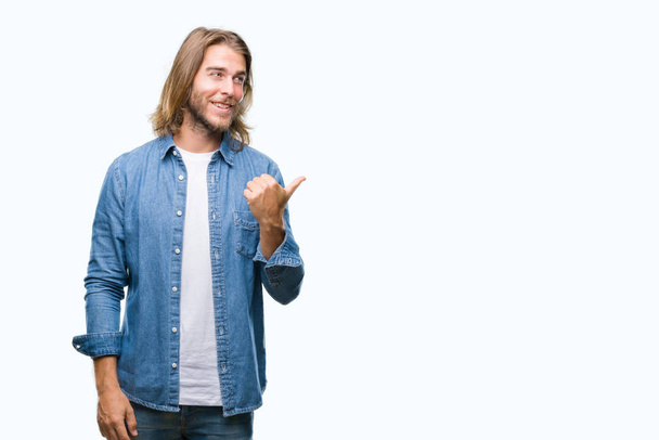 Young handsome man with long hair over isolated background smiling with happy face looking and pointing to the side with thumb up. - Photo, Image