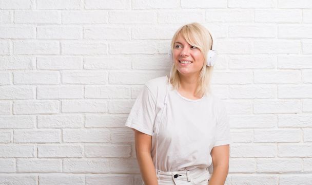 Mulher caucasiana adulta sobre parede de tijolo branco usando fones de ouvido olhando para o lado com sorriso no rosto, expressão natural. Rindo confiante
. - Foto, Imagem