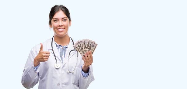 Genç İspanyol doktor kadın holding dolar yapıyor ok işareti, kocaman bir gülümseme ile mutlu parmak ile mükemmel işaret parmak - Fotoğraf, Görsel
