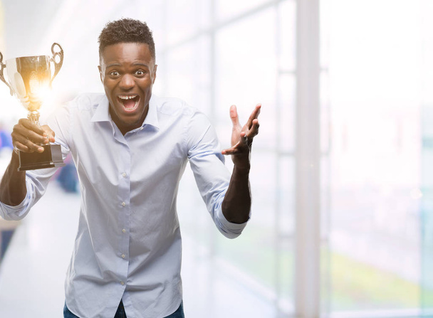 Joven hombre afroamericano sosteniendo trofeo muy feliz y emocionado, expresión ganadora celebrando victoria gritando con gran sonrisa y levantando las manos
 - Foto, imagen