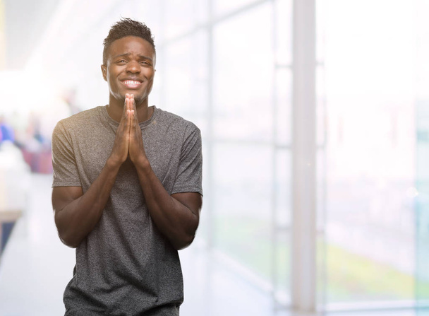 junger afrikanisch-amerikanischer Mann in grauem T-Shirt betet mit den Händen zusammen und bittet um Vergebung, lächelt selbstbewusst. - Foto, Bild