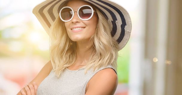 Mladá žena nosí sluneční brýle a letní klobouk s překřížením rukou a spokojené smíchy přirozený úsměv - Fotografie, Obrázek
