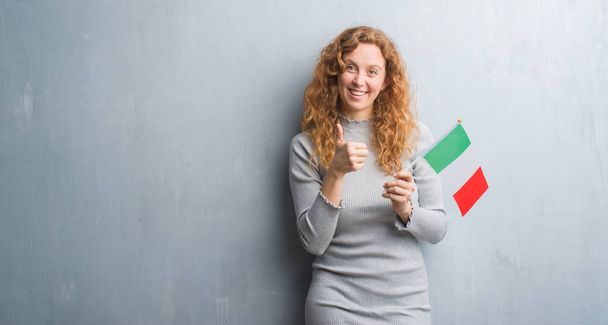Mulher ruiva jovem sobre parede grunge cinza segurando bandeira da Itália feliz com grande sorriso fazendo sinal ok, polegar para cima com os dedos, excelente sinal
 - Foto, Imagem