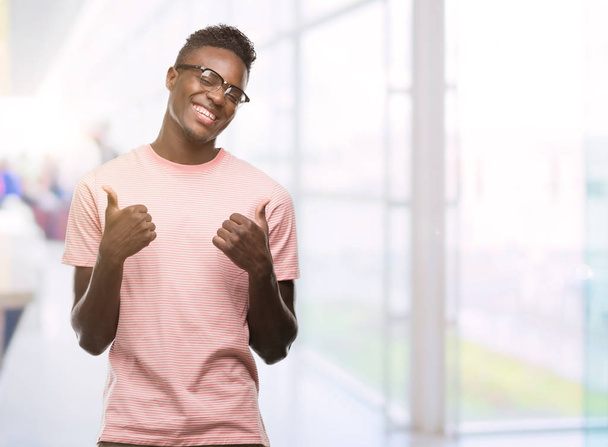 手で、肯定的なジェスチャーをしているピンクの t シャツ成功記号を身に着けている若いアフリカ系アメリカ人は親指を笑顔と幸せです。勝者ジェスチャー晴れやかな表情でカメラを見てください。. - 写真・画像