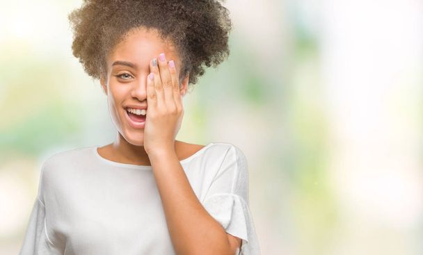 Nuori afro amerikkalainen nainen yli eristetty tausta peittää toisen silmän käsin luottavainen hymy kasvoilla ja yllätys tunteita
. - Valokuva, kuva