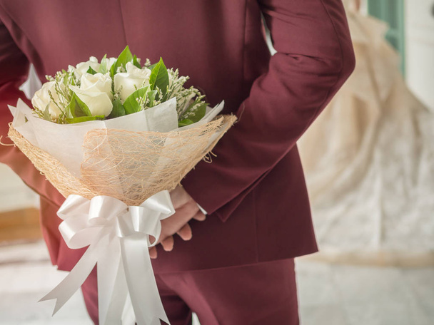 Groom en costume de mariage rouge ou bordeaux cachant bouquet blanc belle fleur de rose avec ruban blanc derrière son dos
. - Photo, image