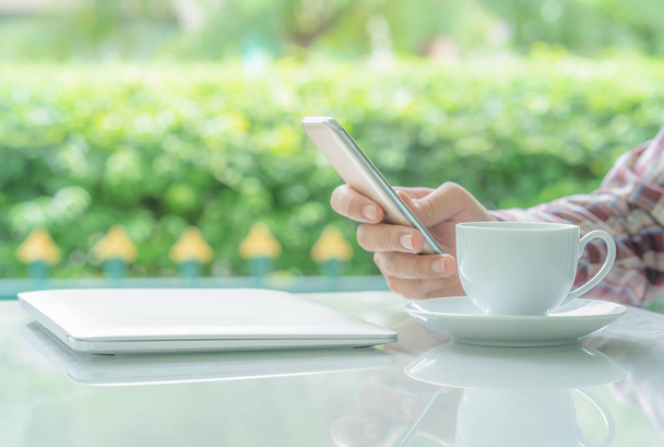 Чоловічі руки тримають смартфон і чашку кави на столі зовні з розмитим зеленим рослинним фоном
 - Фото, зображення