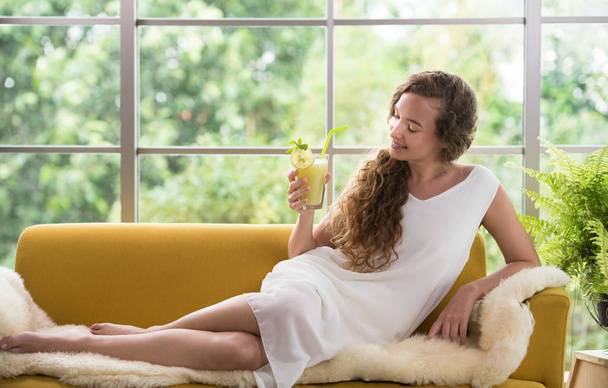 Jovem mulher saudável deitada em um sofá segurando um copo de suco de frutas parecendo relaxado e confortável
 - Foto, Imagem