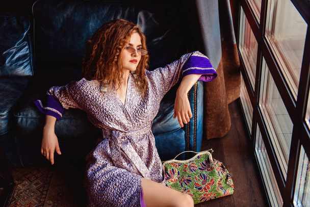 beautiful woman with curly hair in stylish eyeglasses and dress sitting on floor while leaning on sofa near floor window - Foto, afbeelding