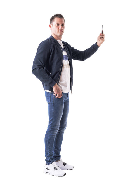 Side view of young man taking photos with mobile phone and looking at camera. Full body isolated on white background. - Foto, Bild