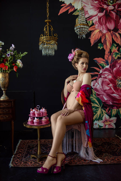 attractive blonde pregnant woman sitting on chair near cupcakes on table in dark decorative floral room  - Фото, зображення