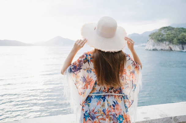 Back view of a young beautiful woman holding her straw hat at the beach and looking to the horizon. - Foto, imagen