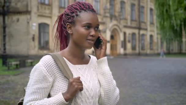 Young charming african female student talking on phone and holding backpack, standing in park near university - Záběry, video