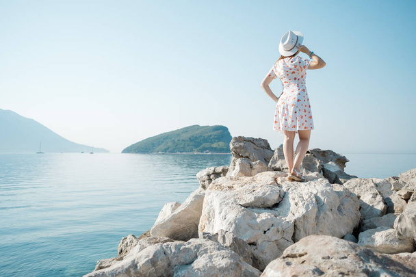 Zadní pohled na mladé krásné ženy drží její bílý klobouk na pláži a při pohledu na obzoru - Fotografie, Obrázek