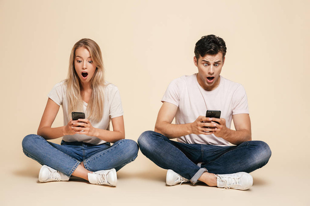 Portrait of a shocked young couple sitting together over beige background, using mobile phones - Foto, Imagem